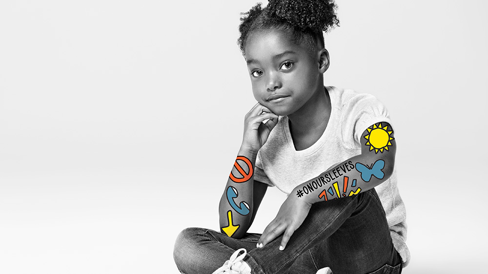 una niña afroamericana sentada de piernas cruzadas, símbolos coloridos #onoursleeves marcan sus brazos.