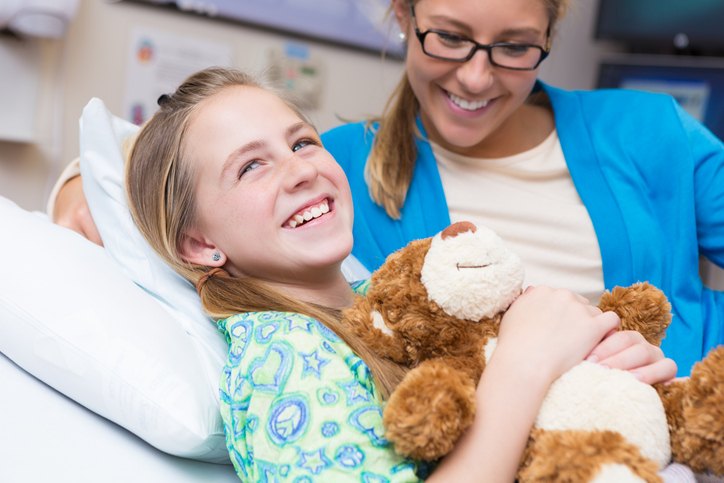 girl holding a teddy bear while she's in a hospital bed, her mother beside her.